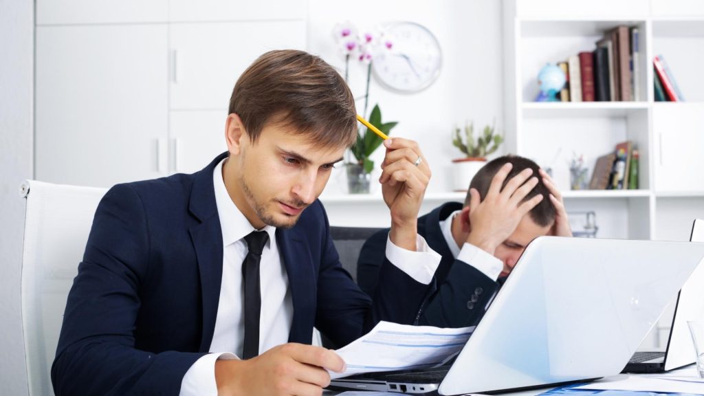 Dos hombres en trajes sentados en un escritorio, trabajando en laptops.