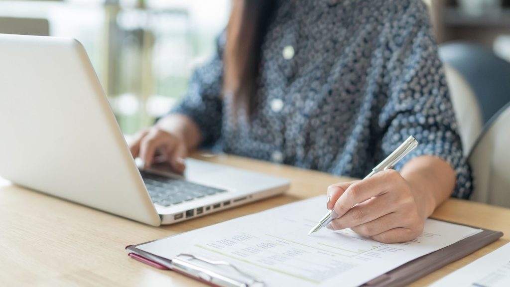 Mujer tomando notas en papel con un bolígrafo, con un laptop abierto frente a ella.
